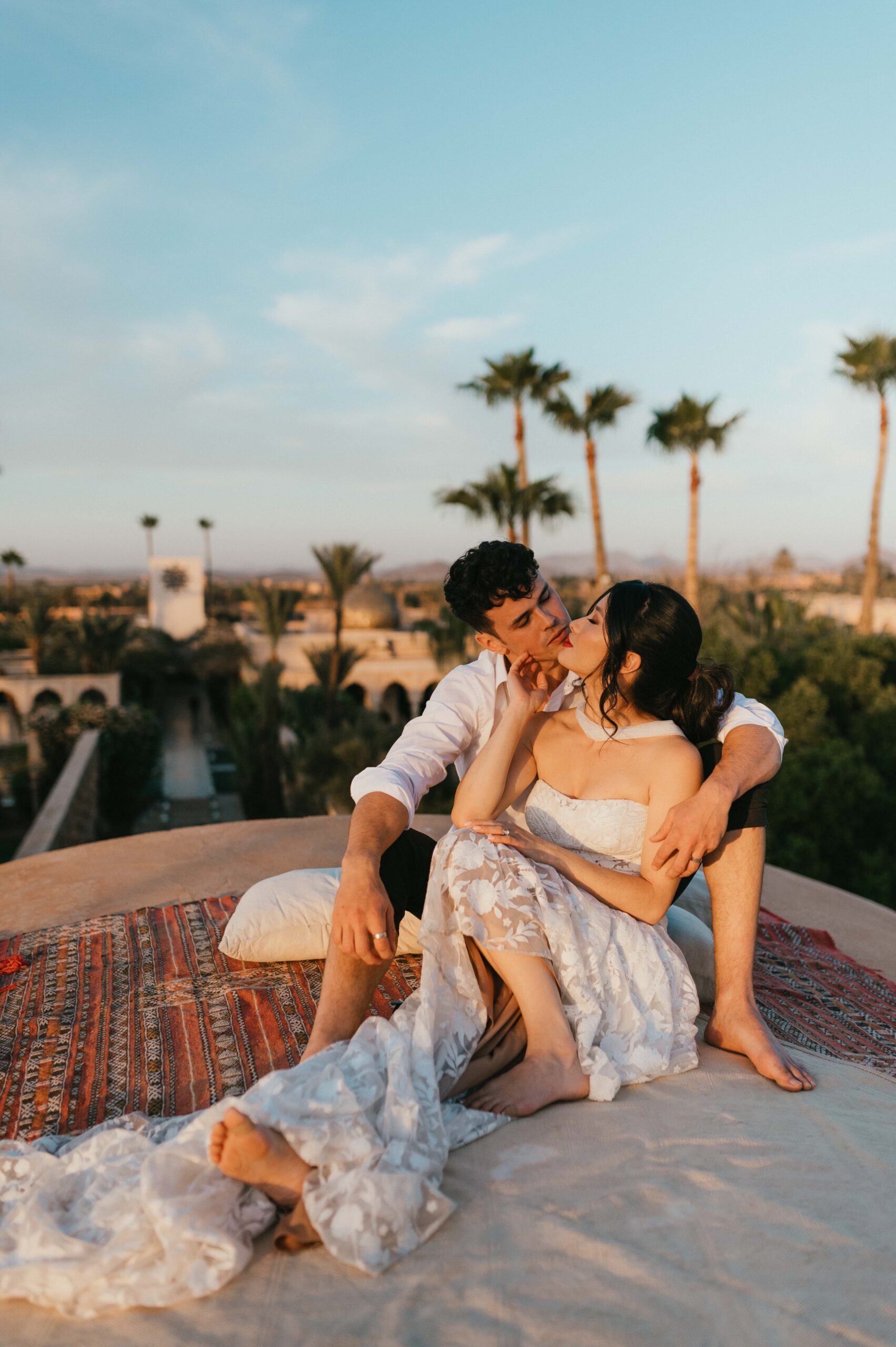 A couple sits together on the dome rooftop of the Palais, with panoramic views of the Marrakech skyline behind them. The bride, Moira, in her Rue de Seine wedding dress, rests comfortably beside her partner, sharing a quiet, intimate moment. The soft breeze and the stunning backdrop of the city below create a serene and romantic atmosphere.