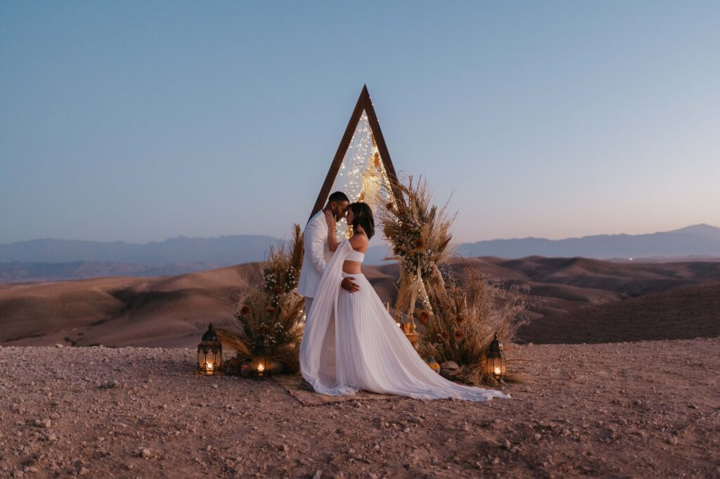 Megan and James exchanging vows in the Marrakech desert, with a beautifully designed setup of Moroccan lanterns, Berber rugs, and soft fairy lights, thanks to the collaboration of La Perle Events Marrakech Wedding Planner, The Bloom Room Marrakech, and Chabichic Morocco.
