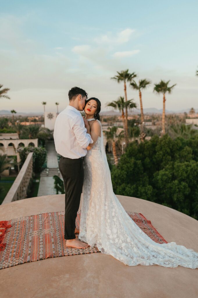 A couple stands on the dome rooftop of the Palais, sharing a tender kiss as they overlook the breathtaking Marrakech skyline. The bride, Moira, in her Rue de Seine wedding dress, wraps her arms around her partner, their love reflected in the moment. The expansive views and the golden light of the setting sun add to the romantic, unforgettable ambiance of their destination wedding.