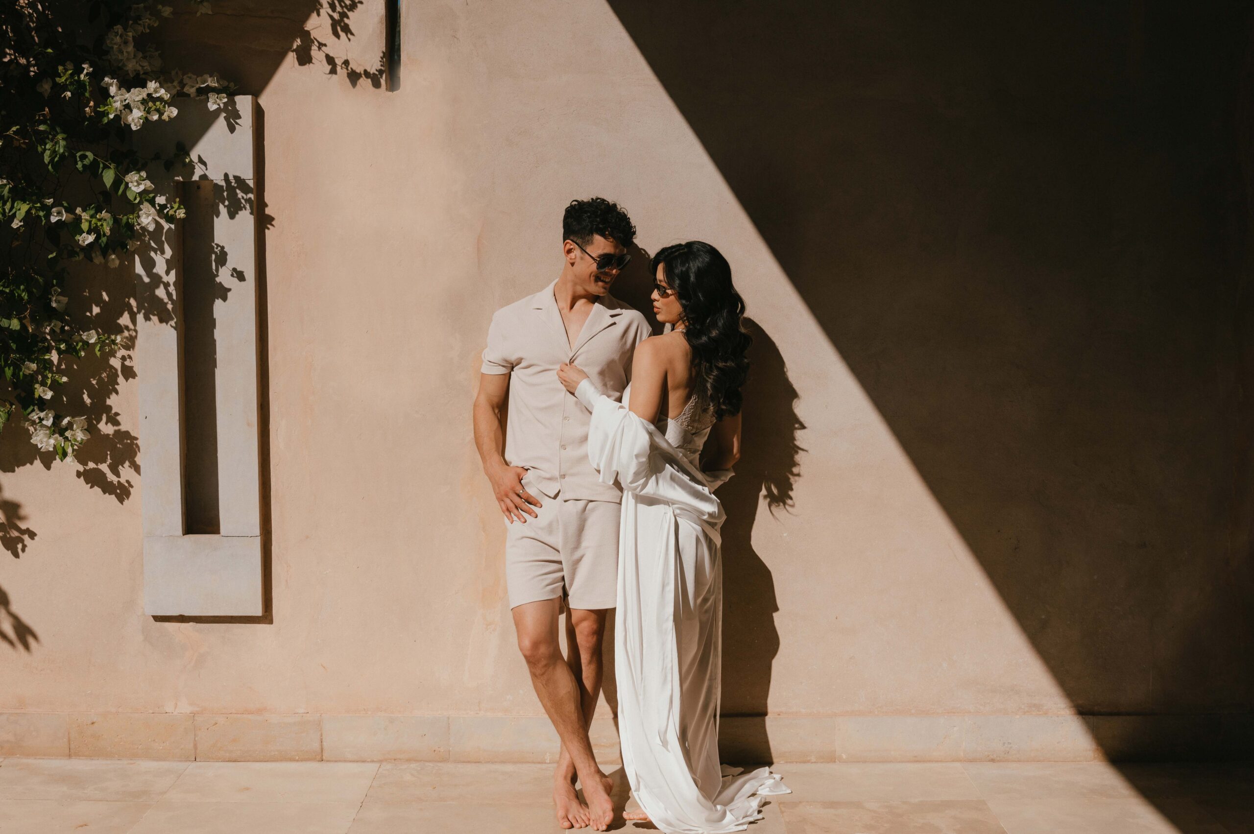 A stylish couple in soft neutral tones, embracing against a sunlit wall in Marrakech, exuding effortless romance and intimacy.