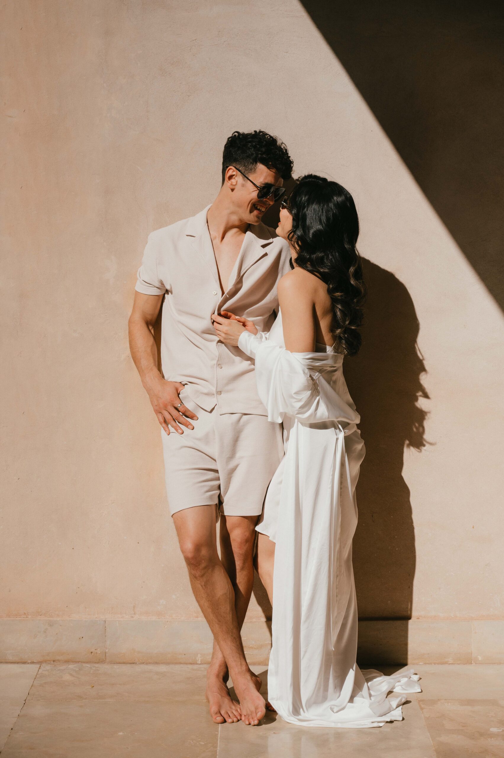 A stylish couple leans into each other against a sunlit Moroccan wall, their effortless connection highlighted by soft shadows and warm tones, dressed in elegant yet relaxed wedding-inspired attire.