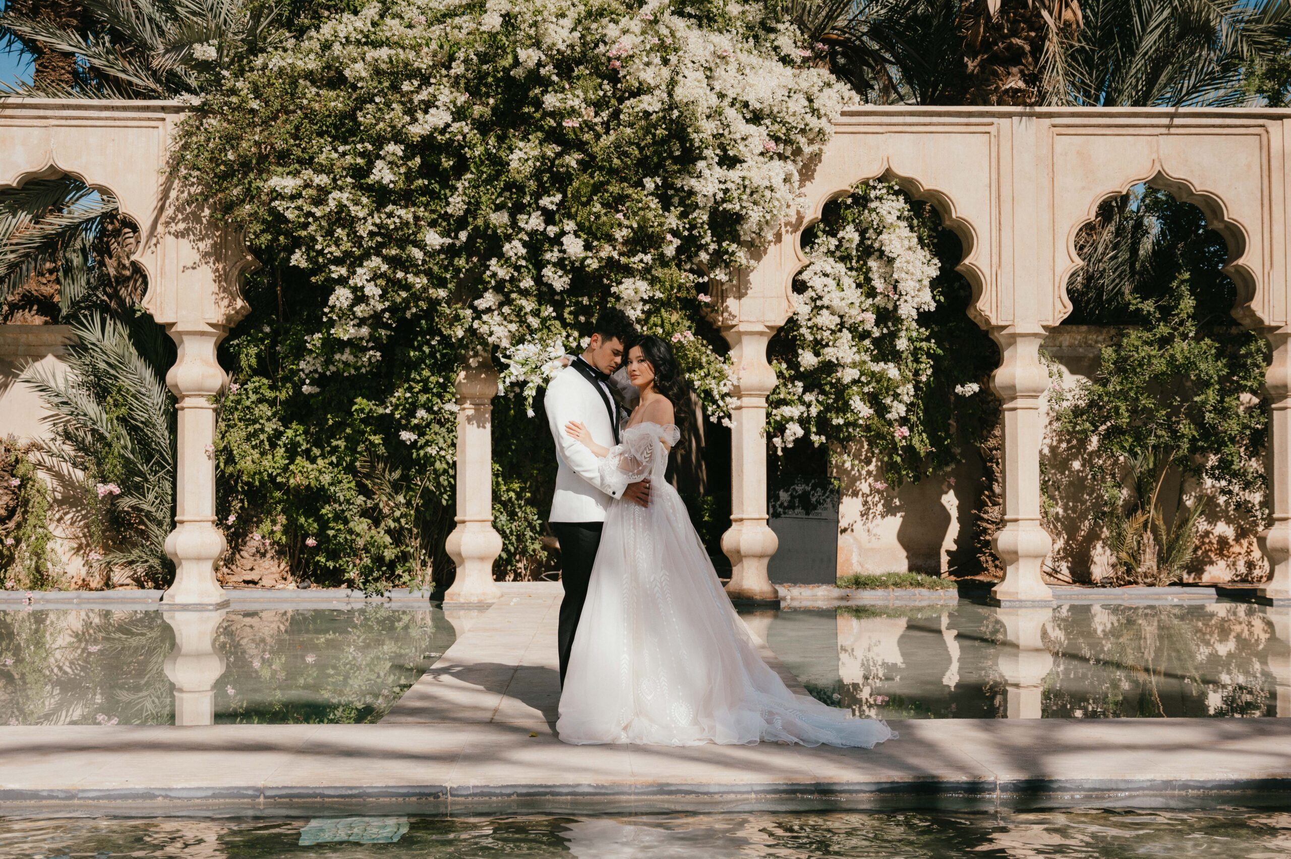 A romantic couple’s portrait by the reflective pool at Palais Namaskar, Marrakech, with the water mirroring the stunning Moroccan architecture and golden light.