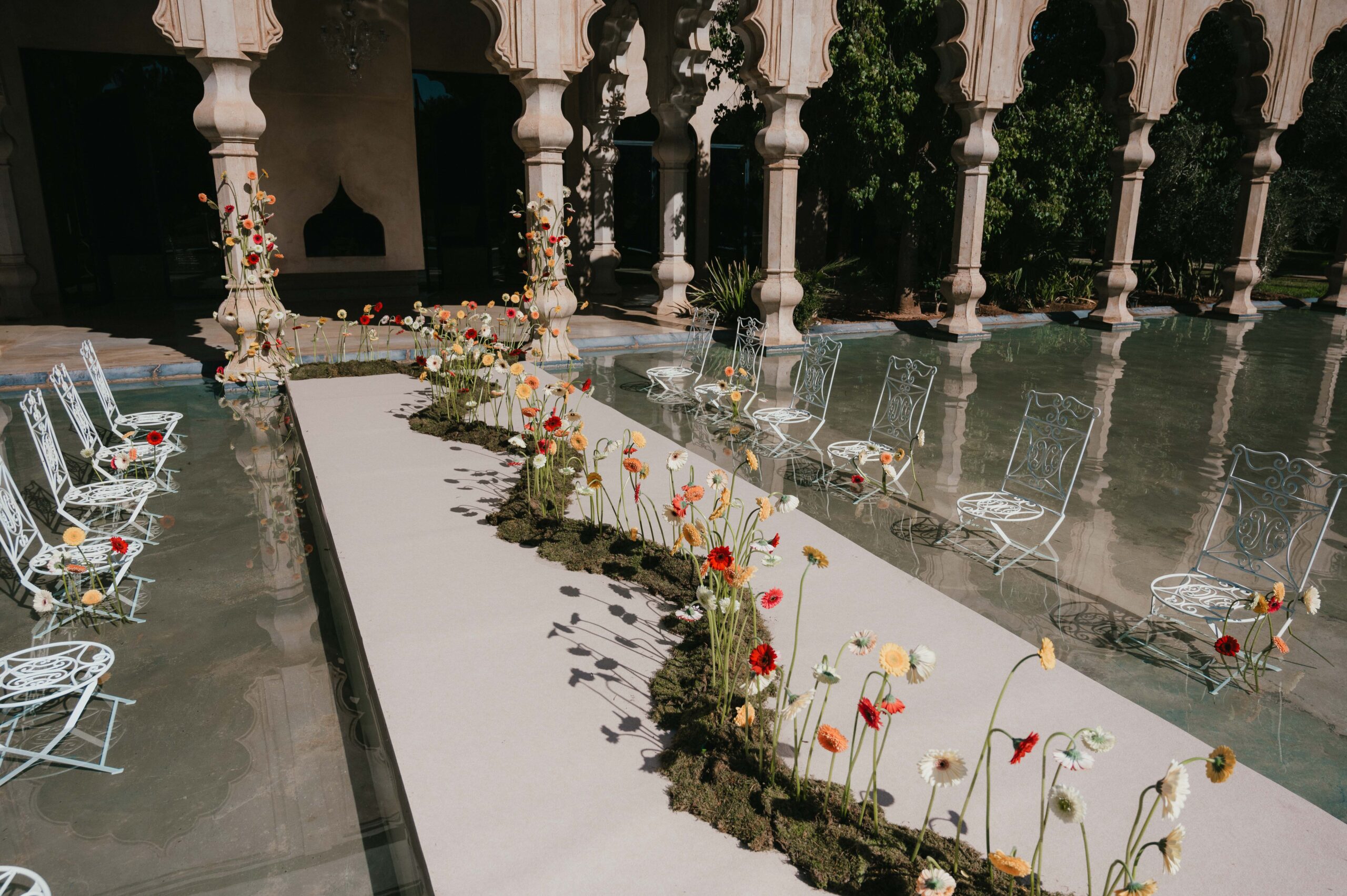 A stunning wedding ceremony setup at Palais Namaskar, Marrakech, featuring white chairs arranged in shallow water, reflecting the golden hues of the Moroccan sun and the venue’s elegant architecture.