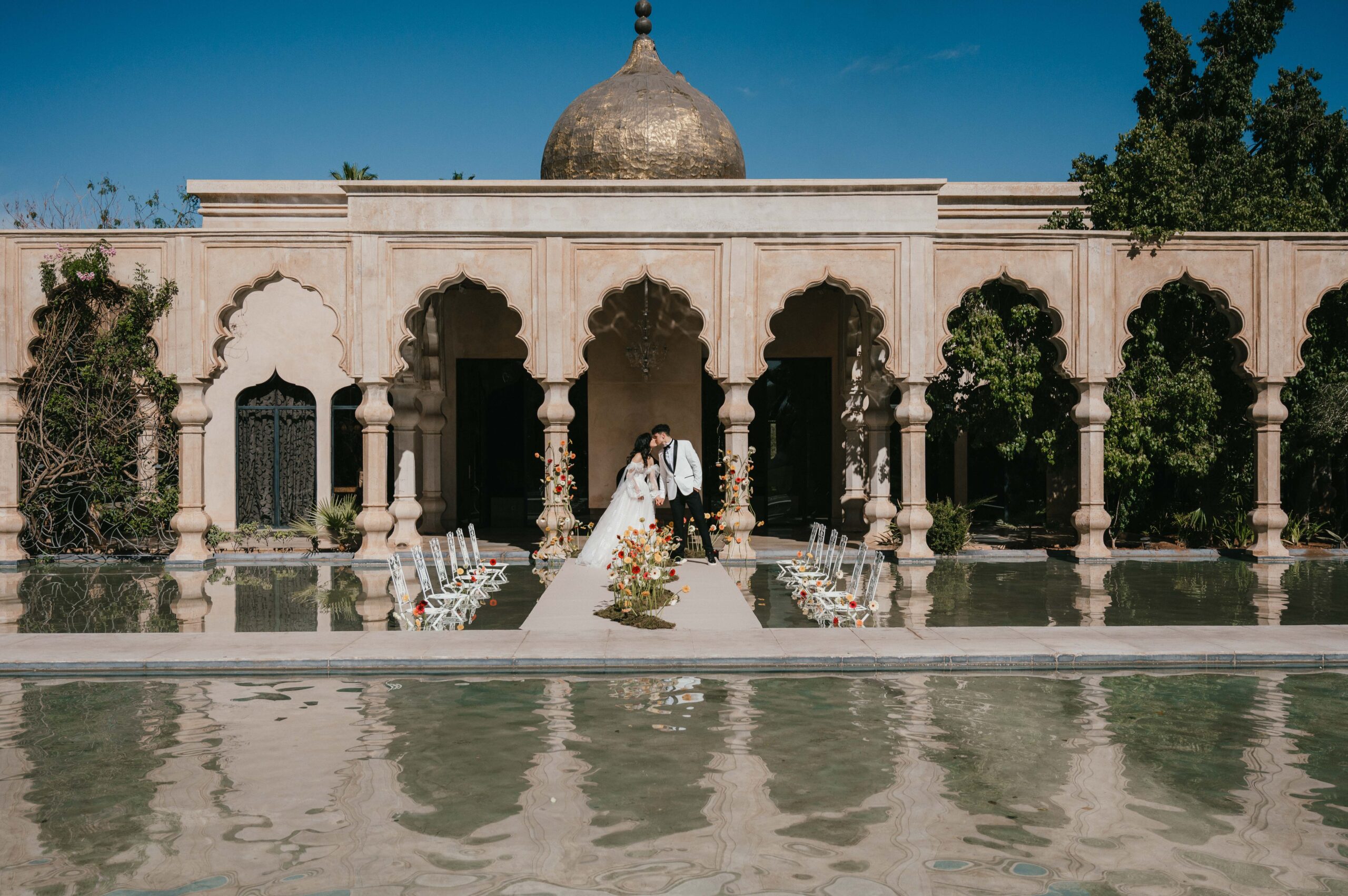 A breathtaking wedding ceremony setup at Palais Namaskar, Marrakech, with white chairs placed in the shallow water, reflecting the golden light and stunning Moroccan architecture.