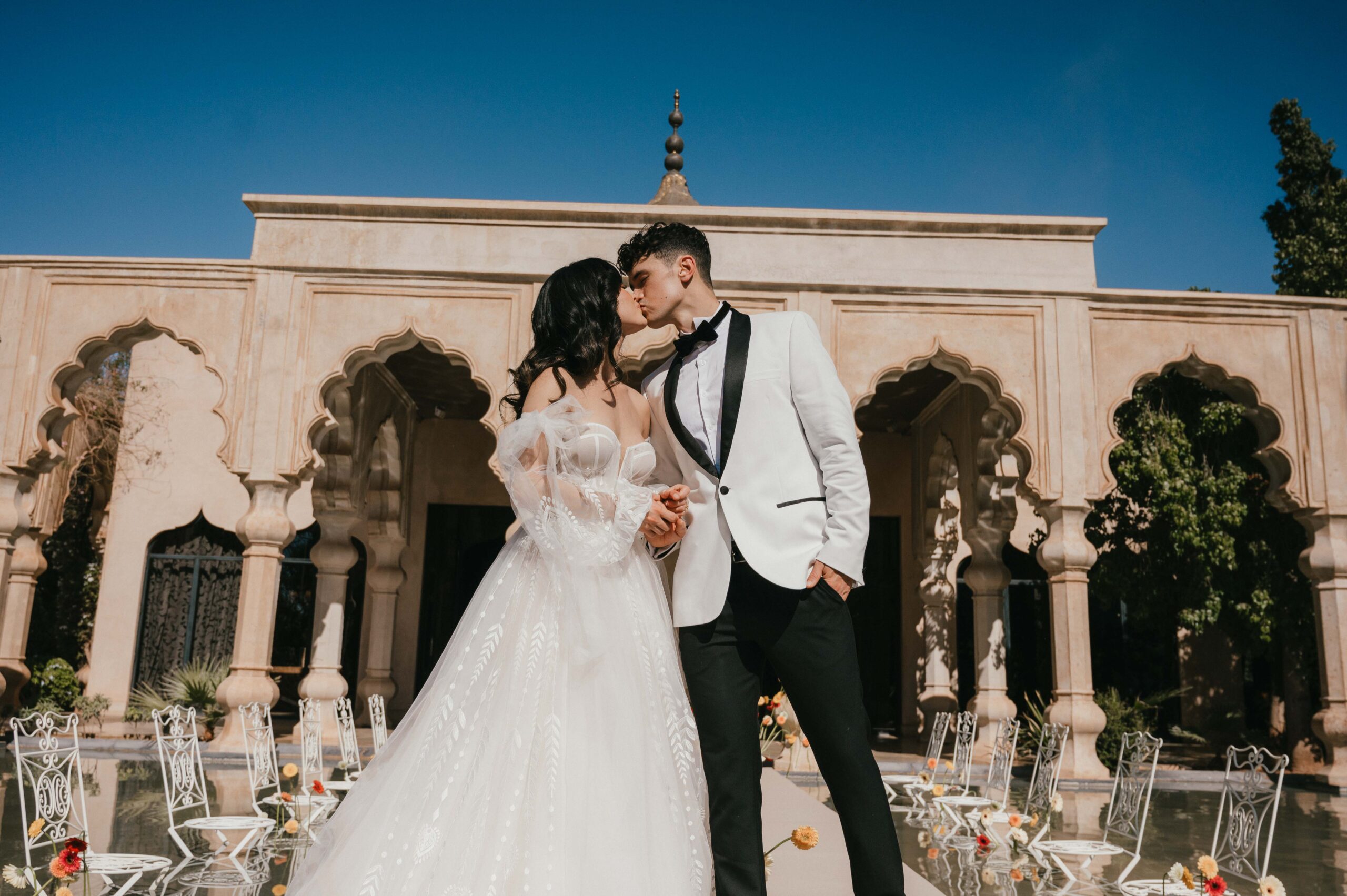 Bride and groom share a romantic first kiss at their Palais Namaskar wedding ceremony, with the bride wearing an off-the-shoulder gown from the Oasis Collection by Rich Bridal, framed by intricate Moroccan arches and elegant floral décor.