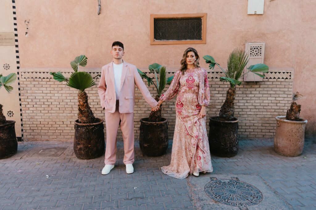 Paul, sporting a stylish pink suit, and Lisa, radiant in her Rue de Seine bridal gown, hold hands as they stand beside potted palm trees against a striking pink wall in Marrakech.
