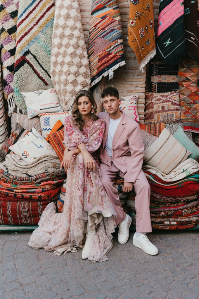 Alt text: Paul and Lisa, dressed in a Rue de Seine bridal gown, sit together on vibrant carpets and cushions in the bustling Marrakech souk, surrounded by colourful textiles and traditional decor.