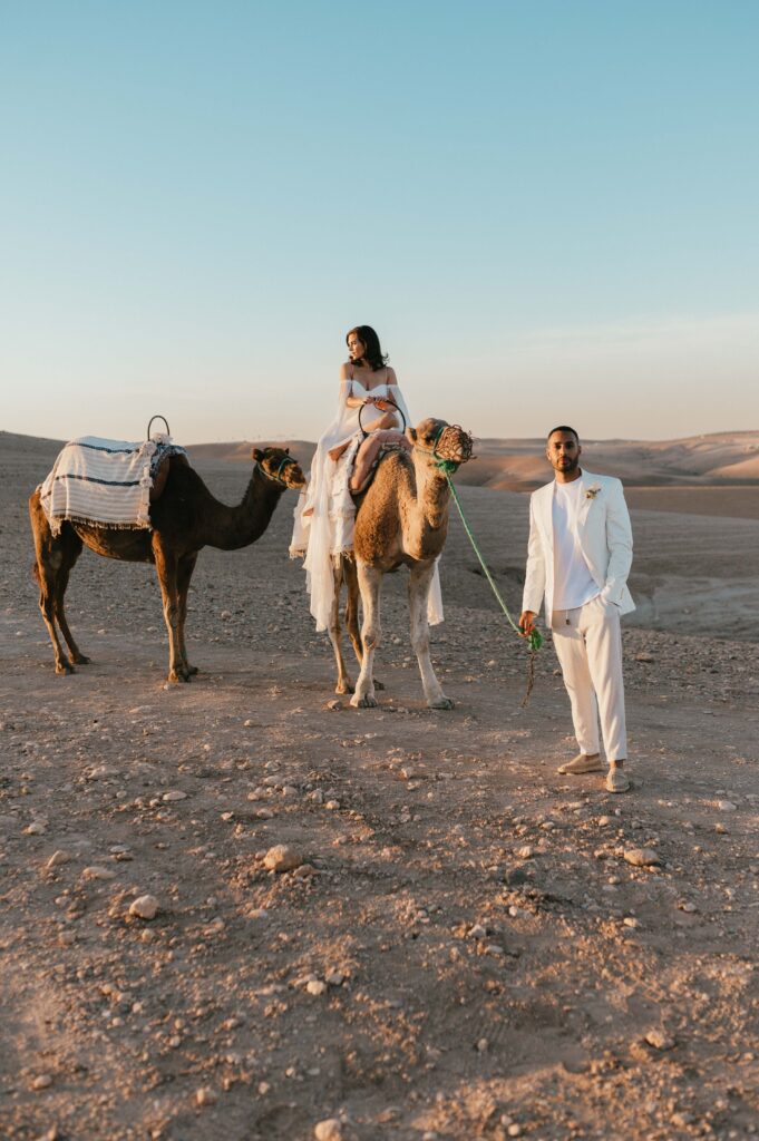 Megan riding a camel while James pulls the two camels, with the Moroccan desert landscape stretching out behind them.