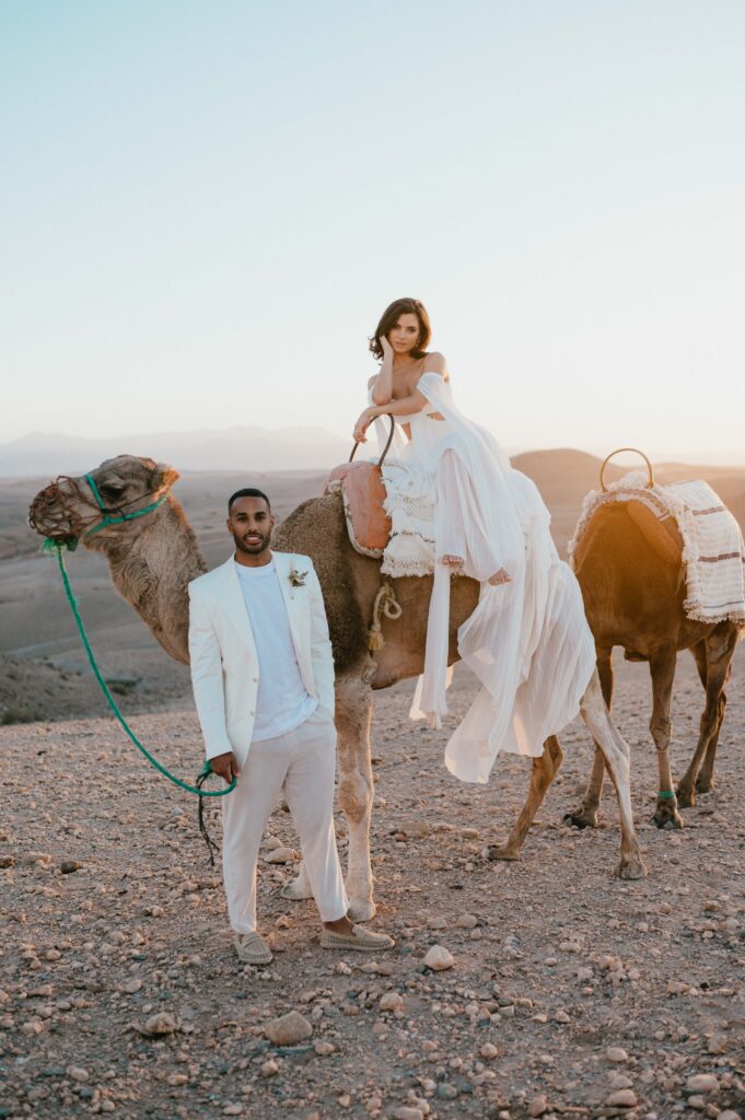Megan, dressed in a Rue de Seine gown with floating sleeves, riding a camel in the Agafay desert at sunrise, bathed in warm golden light.