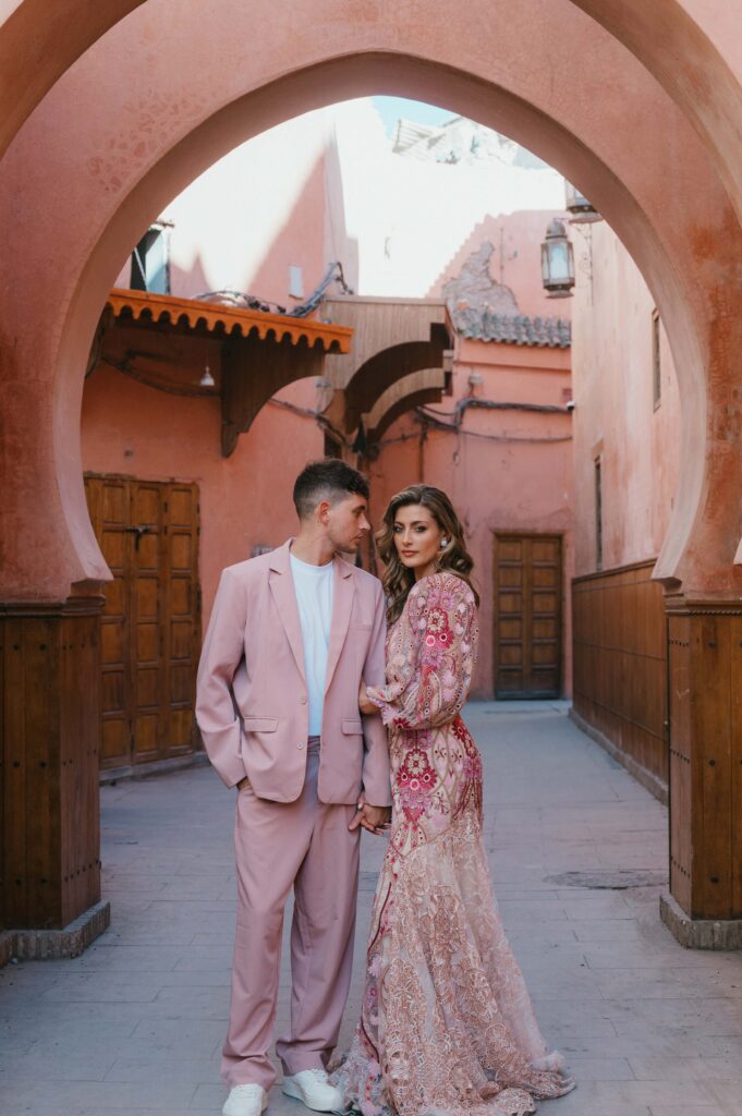 Paul and Lisa stand beneath a vibrant pink doorway arch in the bustling Marrakech Medina souk, surrounded by traditional architecture and lively market energy.