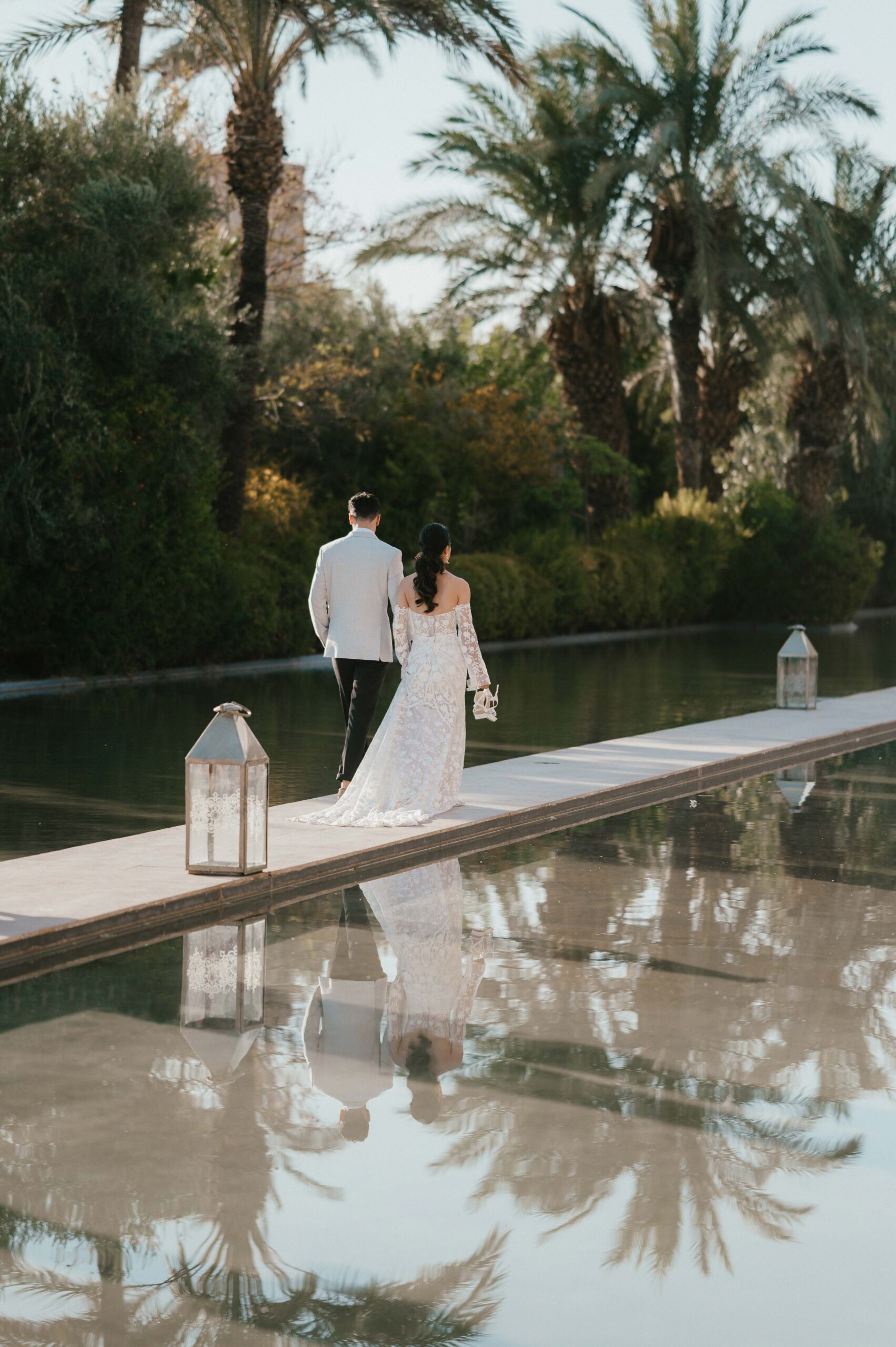 A couple walks hand in hand along a serene pathway that stretches across calm water, their reflections beautifully mirrored on the surface. The bride, Moira, in her Rue de Seine wedding dress, gently steps alongside her partner, creating a moment of pure tranquility. The peaceful surroundings and the perfect reflection add a magical, ethereal quality to their destination wedding experience.