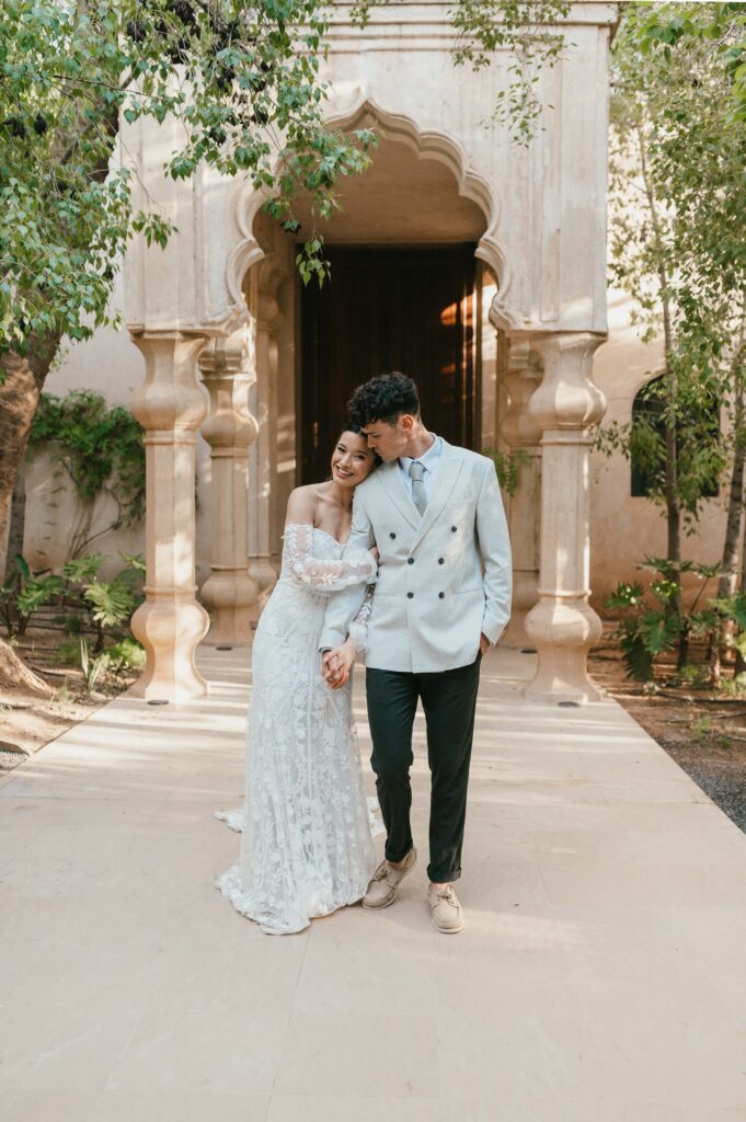 A couple stands in a beautiful garden by a Moroccan-style door. The bride, Moira, is wearing a Rue de Seine wedding dress with intricate lace details, holding her partner's hand while they share a tender moment. The lush greenery and the unique door frame add to the romantic atmosphere of the scene.
